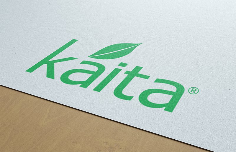 Logotipo de Kaita, con tipografía verde y una hoja, sobre fondo blanco y superficie de madera. Reflexiona un enfoque ecológico.