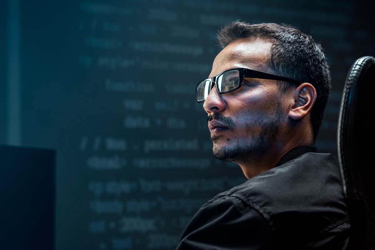 Hombre con gafas mirando atentamente a la pantalla, rodeado de código en un entorno de trabajo tecnológico.