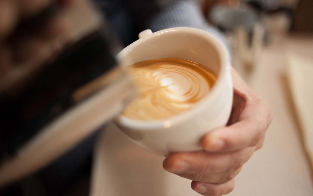 Taza de café con un diseño artístico en la superficie, mientras una mano sirve la bebida en un ambiente acogedor.