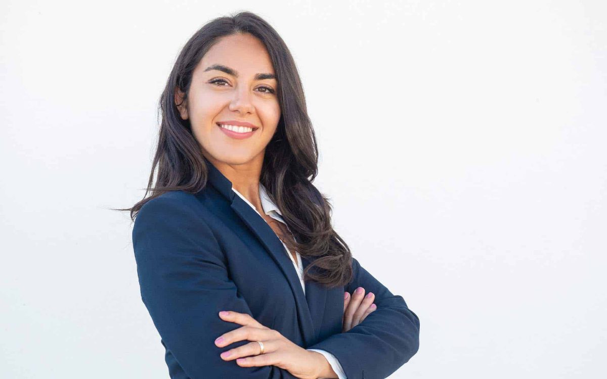 Mujer sonriente con traje formal, de brazos cruzados, sobre fondo blanco, transmitiendo confianza y profesionalismo.