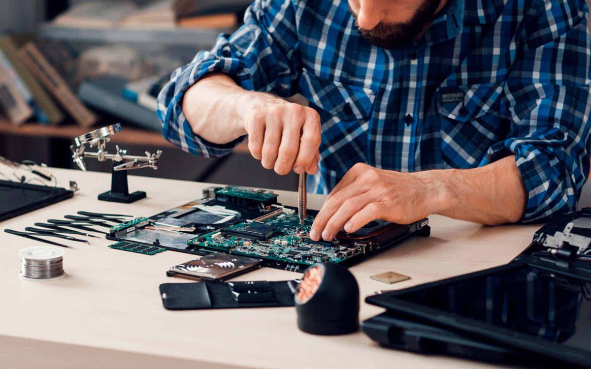 Técnico repara un ordenador portátil, usando herramientas en una mesa con componentes electrónicos y equipos de trabajo.