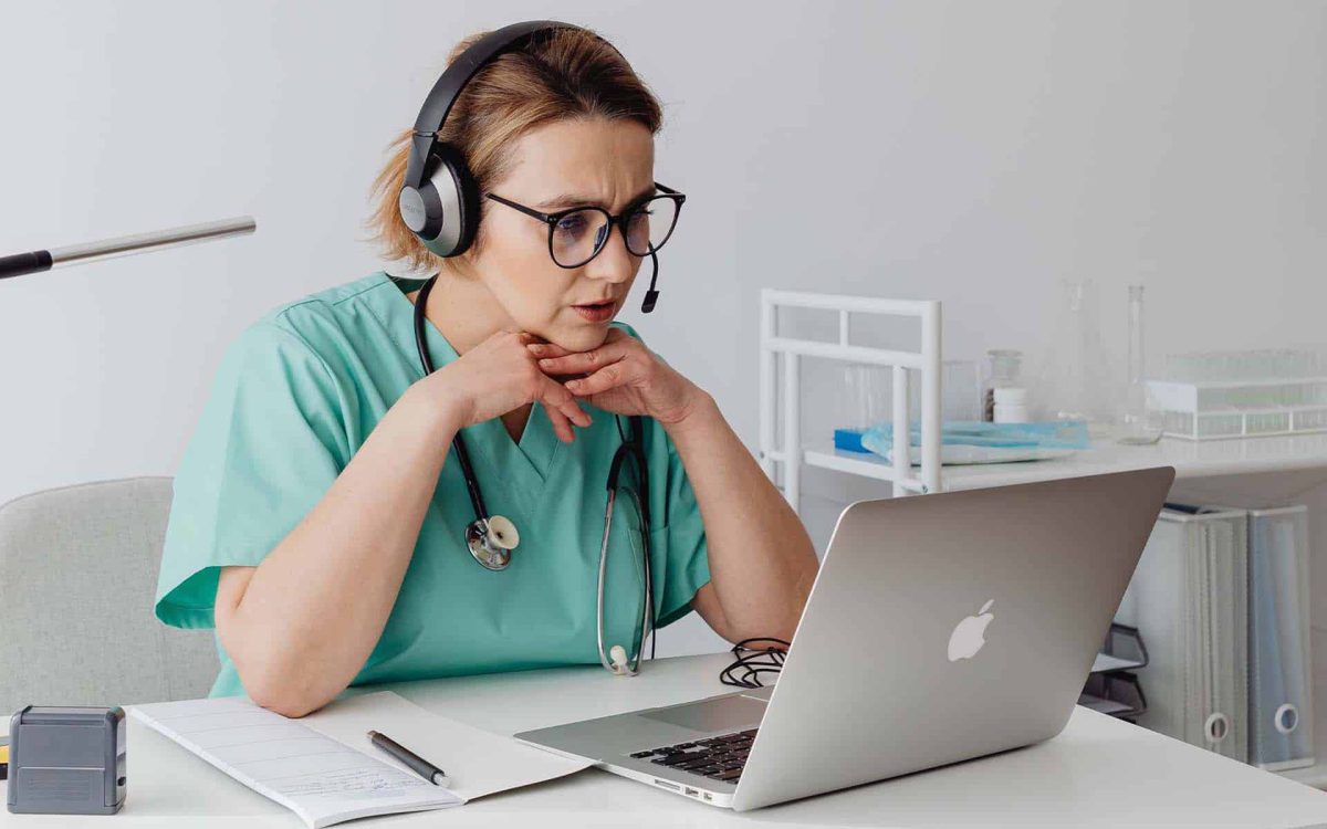 Mujer en uniforme médico, usando auriculares, concentrada en una consulta virtual frente a su laptop.