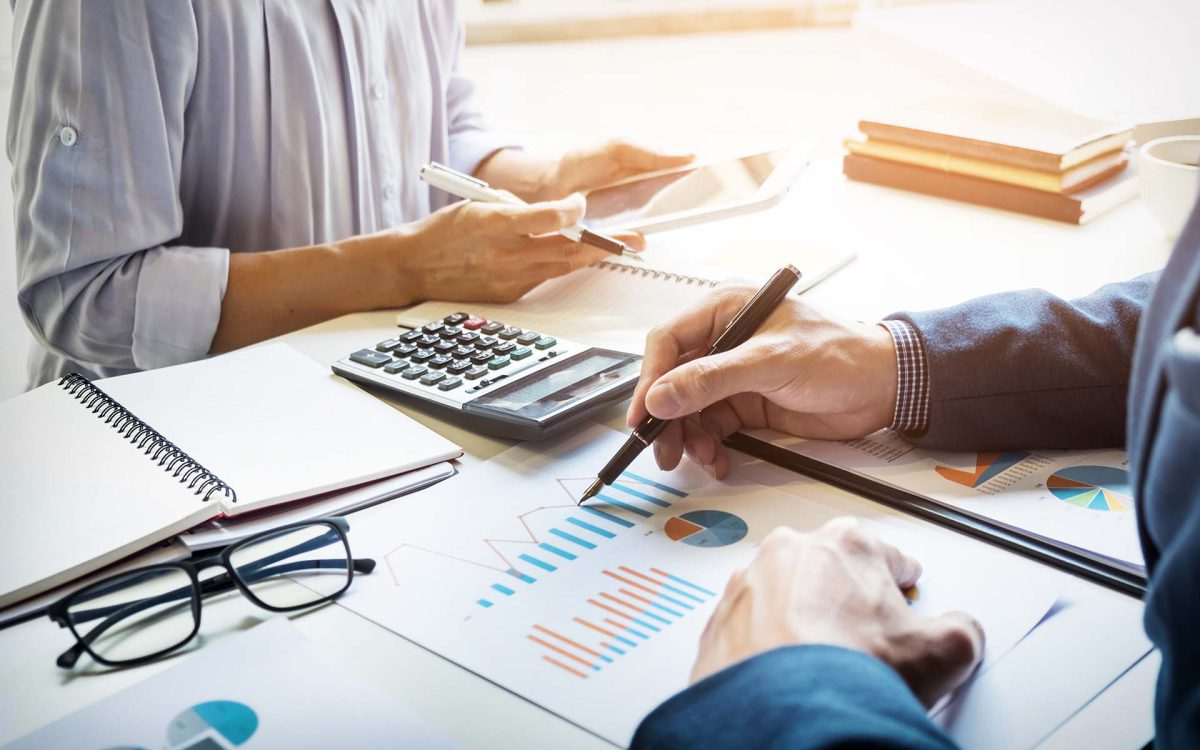 Personas trabajando en informes financieros, con gráficos, calculadora y tablet sobre la mesa. Ambiente de oficina.