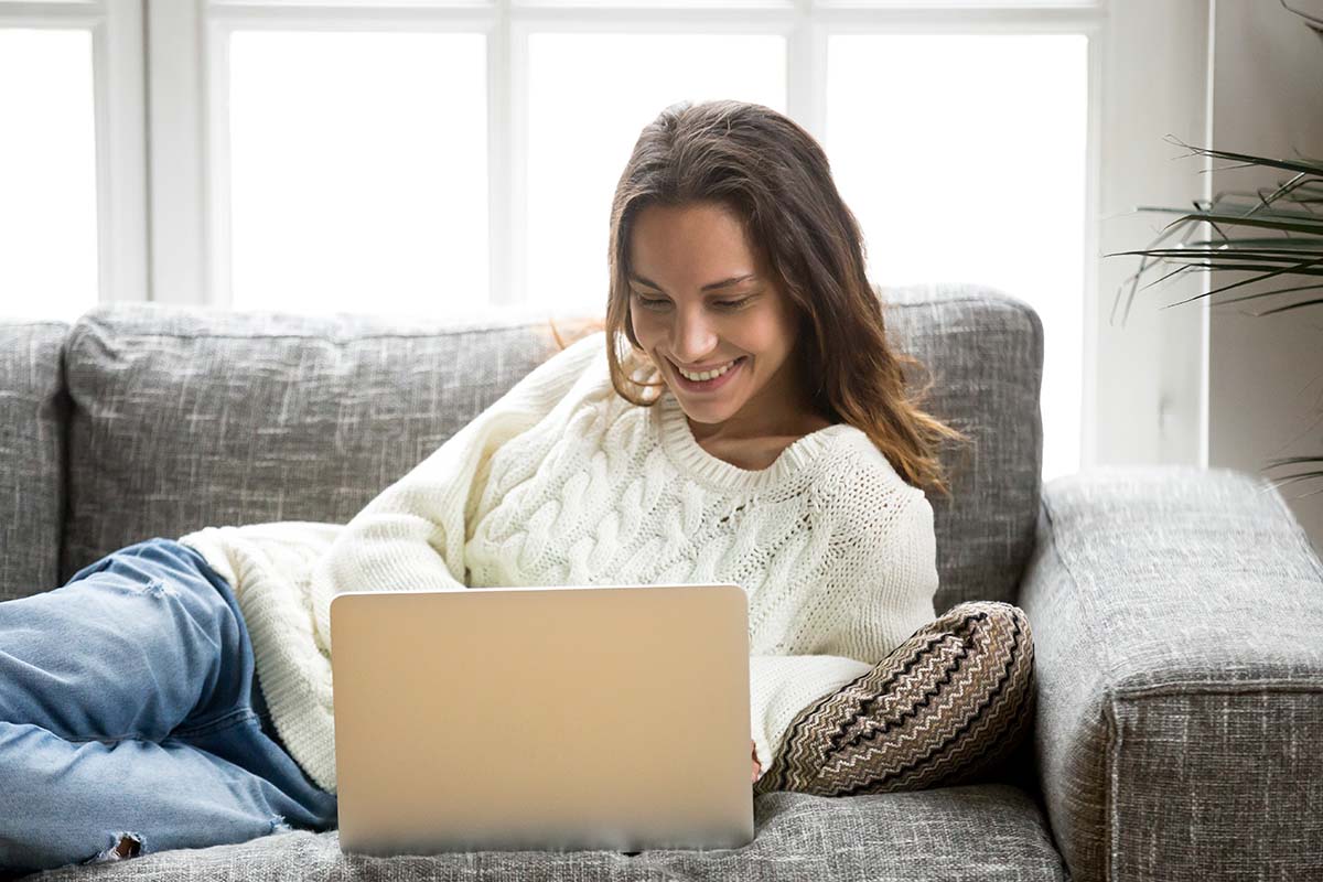 Mujer sonriente usando una laptop en un sofá cómodo, disfrutando de un momento relajante en casa.