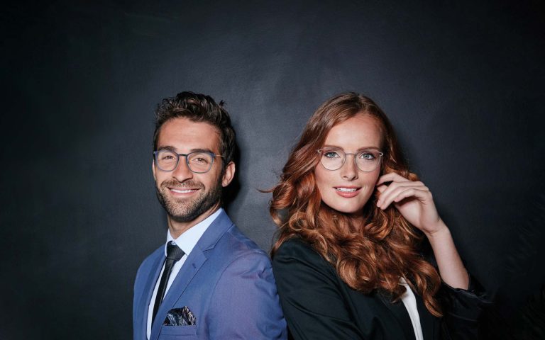 Pareja posando con gafas elegantes, sonriendo, en un fondo oscuro que resalta sus atuendos formales y el ambiente profesional.