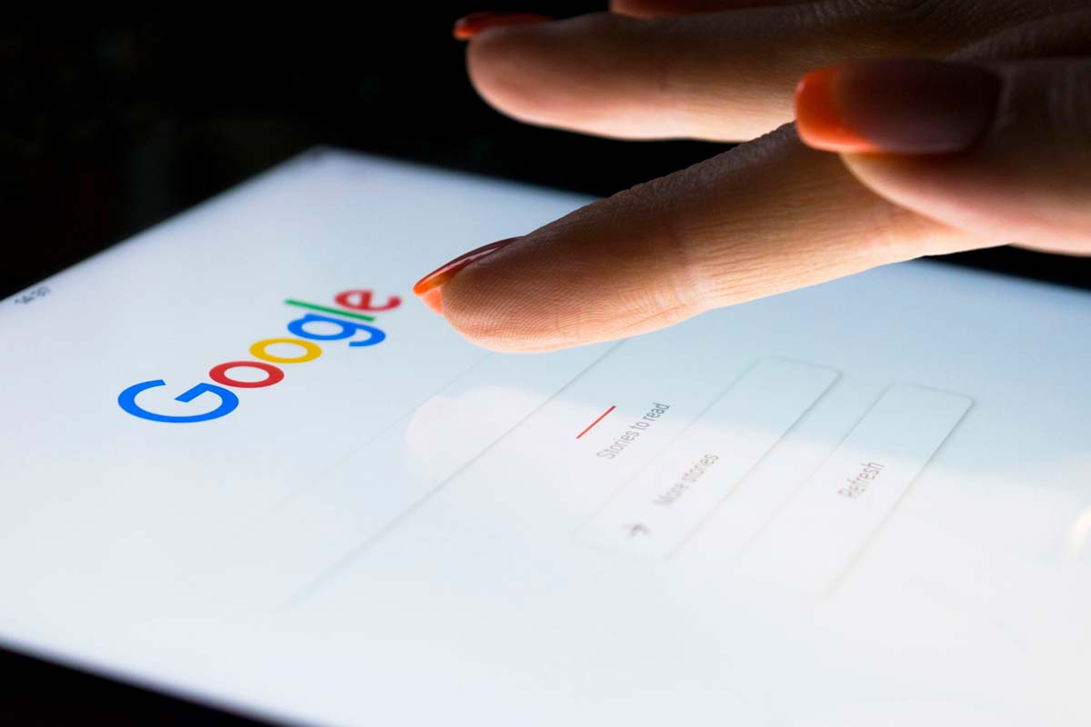 Mano con uñas pintadas acercándose a la pantalla de un dispositivo mostrando el logo de Google y su barra de búsqueda.
