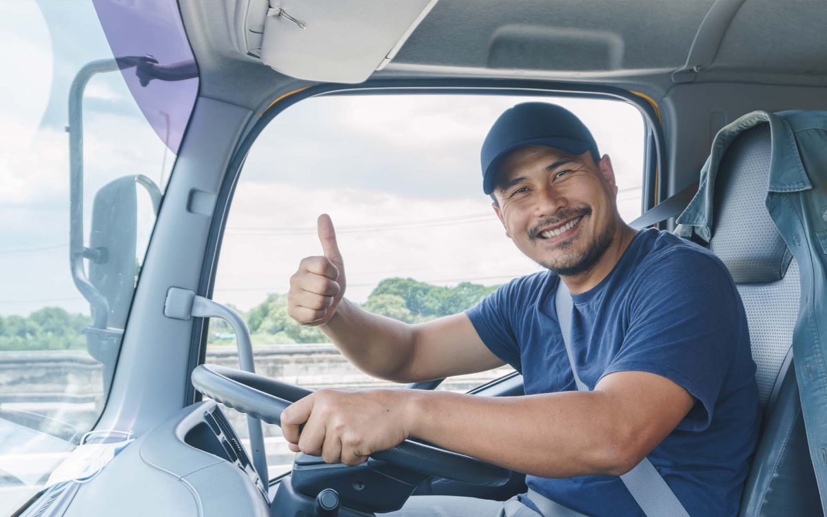 Conductor sonriente de camión mostrando el pulgar hacia arriba, sentado al volante en un entorno natural.