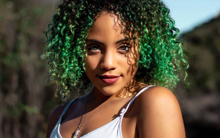 Mujer con cabello rizado y verde, mirada intensa, fondo natural iluminado por el sol. Captura de belleza y estilo.