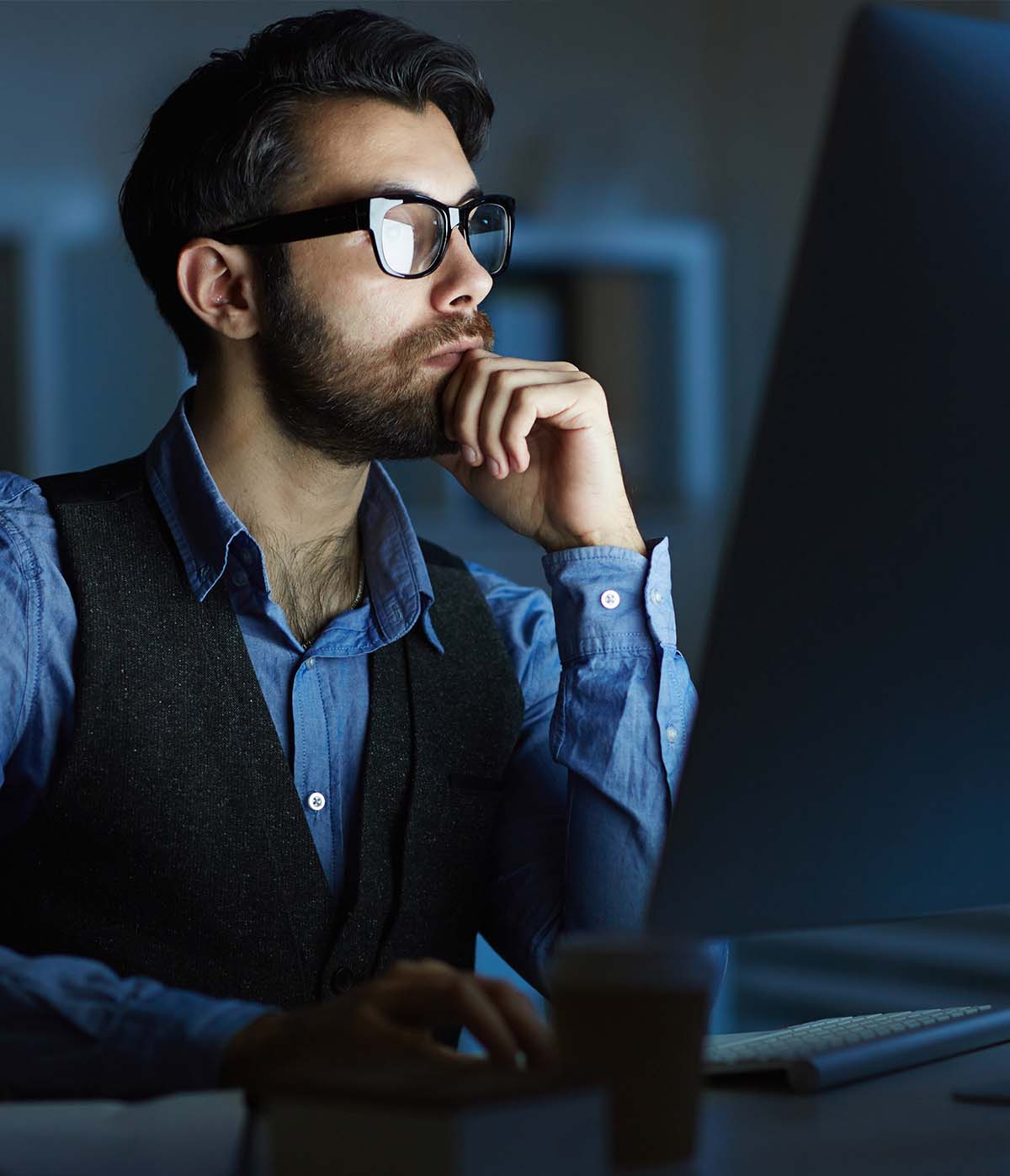 Hombre con gafas pensando frente a una computadora en un ambiente de oficina oscura, mostrando concentración y reflexión.