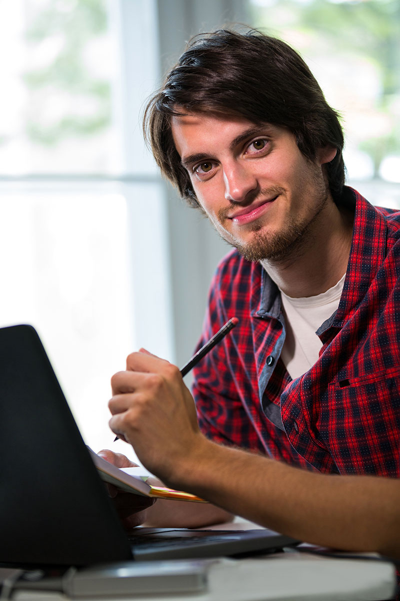 Joven sonriente trabajando en su laptop, tomando notas con un lápiz en un ambiente luminoso y motivador.