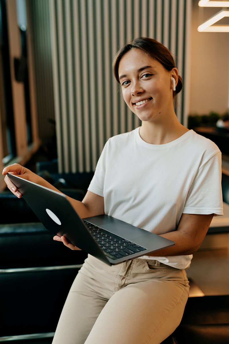 Mujer sonriente con laptop en mano, vistiendo camiseta blanca, sentada en un ambiente moderno y luminoso.