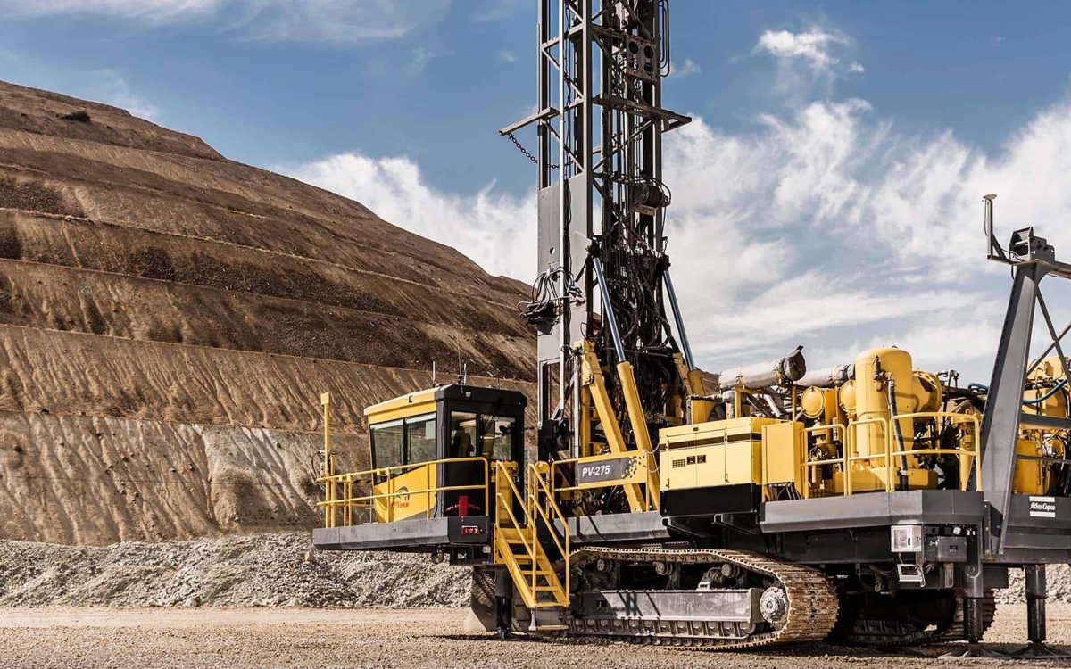 Máquina perforadora amarilla en un sitio de minería con montañas de tierra en el fondo y cielo despejado.