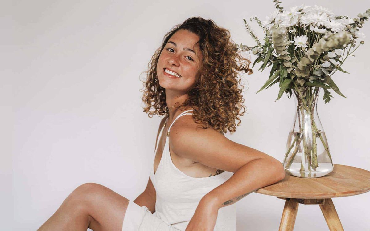 Mujer sonriente con cabello rizado, sentada junto a una mesa con un jarrón de flores en fondo blanco.