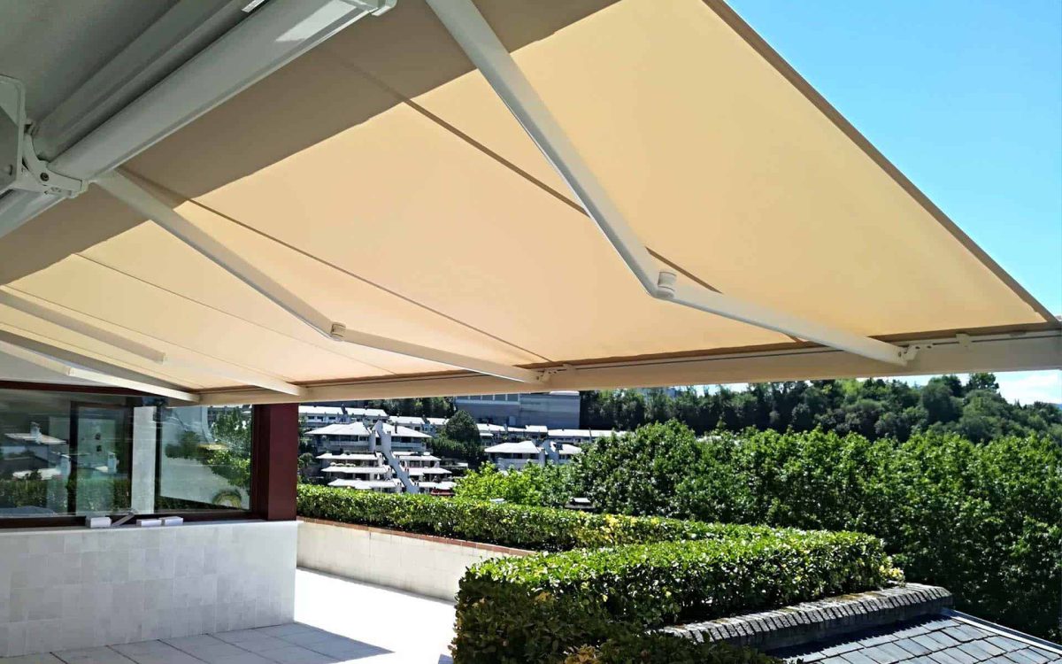 Toldo retráctil beige instalado en terraza, con vistas a edificios y naturaleza en un día soleado.