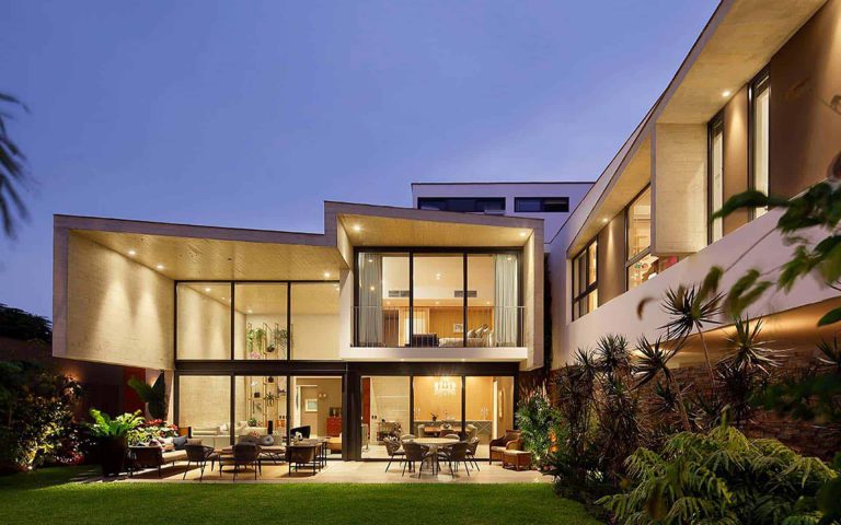 Casa moderna iluminada al anochecer, con amplios ventanales y un jardín bien cuidado. Espacio acogedor y elegante.